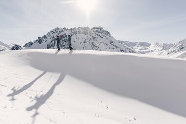 Zwei Skifahrer gehen mit Ski über der Schulter durch tiefen Schnee in Gargellen, lange Schatten im Vordergrund. | © Gargellner Bergbahnen, Daniel Zangerl