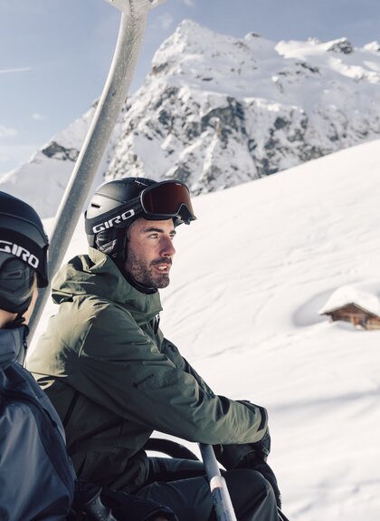 Zwei Skifahrer, eine Frau und ein Mann, sitzen gemeinsam in einem Sessellift und fahren den verschneiten Berg hinauf. Beide tragen Skibekleidung, Helme und Skibrillen. Im Hintergrund erstreckt sich eine tiefverschneite Winterlandschaft mit einem kleinen, fast eingeschneiten Holzhaus und einem imposanten, schneebedeckten Bergmassiv, der Madrisa. Die Stimmung ist ruhig und sonnig, der Himmel klar. | © Gargellner Bergbahnen GmbH & CO KG, Daniel Zangerl