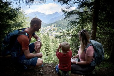 Eine Familie mit Kind sitzt am Waldrand oberhalb von Gargellen und blickt auf das Tal mit Häusern, Wiesen und Bergen im Hintergrund. Die Erwachsenen tragen Rucksäcke, das Kind schaut in die Ferne. | © Montafon Tourismus, Stefan Kothner