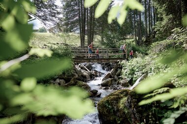 Eine Familie mit zwei Kindern wandert über eine kleine Holzbrücke in einem grünen Bergwald in Gargellen. Unter der Brücke fließt ein klarer Gebirgsbach zwischen Felsen, umgeben von Bäumen und Wiesen. | © Montafon Tourismus, Stefan Kothner