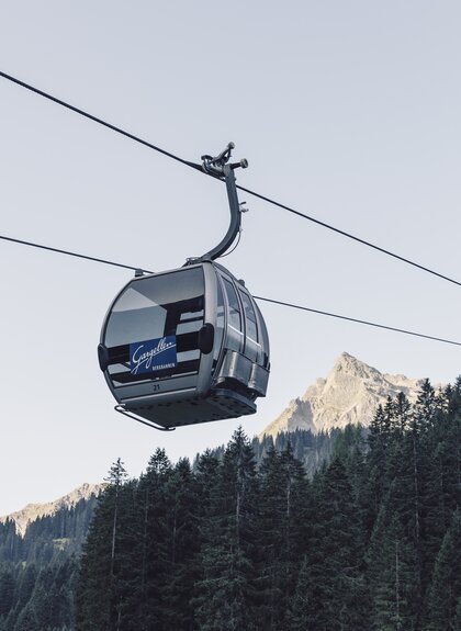 ine silberne Gondel der Gargellner Bergbahnen schwebt über einen dichten Nadelwald in Richtung Bergstation. Im Hintergrund ragt der Gipfel der Madrisa in der Abendsonne auf, während der Himmel klar und hellblau ist. | © Gargellner Bergbahnen GmbH & Co KG, Daniel Zangerl