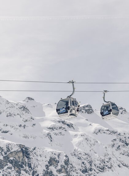 Zwei Gondeln der Schafbergbahn in Gargellen schweben vor einer beeindruckenden, schneebedeckten Bergkulisse. Die modernen Kabinen der Gargellner Bergbahnen heben sich klar vom hellen Winterhimmel ab, während im Hintergrund die majestätischen Gipfel der Montafoner Alpen aufragen. | © Gargellner Bergbahnen GmbH & Co KG, Daniel Zangerl
