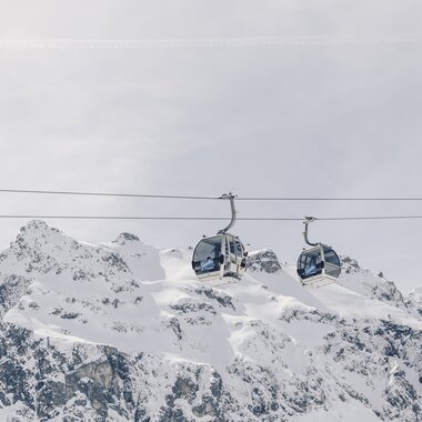 Zwei Gondeln der Schafbergbahn in Gargellen schweben vor einer beeindruckenden, schneebedeckten Bergkulisse. Die modernen Kabinen der Gargellner Bergbahnen heben sich klar vom hellen Winterhimmel ab, während im Hintergrund die majestätischen Gipfel der Montafoner Alpen aufragen. | © Gargellner Bergbahnen GmbH & Co KG, Daniel Zangerl