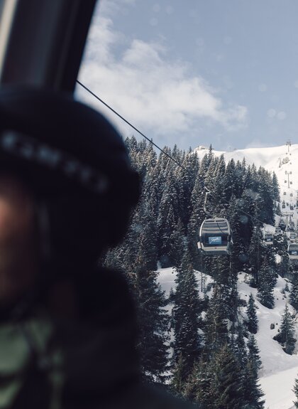 Ein Skifahrer mit Helm und Skibrille sitzt in einer Gondel der Schafbergbahn in Gargellen. Durch das Fenster ist die Seilbahn zu sehen, die über verschneite Wälder und Hänge führt. Im Hintergrund steigen die Gondeln der Gargellner Bergbahnen den schneebedeckten Berg hinauf. | © Gargellner Bergbahnen GmbH & Co KG, Daniel Zangerl