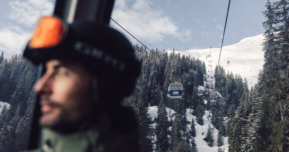 Ein Skifahrer mit Helm und Skibrille sitzt in einer Gondel der Schafbergbahn in Gargellen. Durch das Fenster ist die Seilbahn zu sehen, die über verschneite Wälder und Hänge führt. Im Hintergrund steigen die Gondeln der Gargellner Bergbahnen den schneebedeckten Berg hinauf. | © Gargellner Bergbahnen GmbH & Co KG, Daniel Zangerl