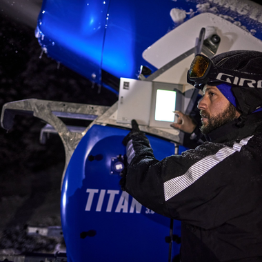 Zwei Mitarbeiter der Gargellner Bergbahnen überwachen nachts eine laufende Schneekanone. Die Maschine sprüht feinen, künstlichen Schnee über die beleuchtete Skipiste, während im Hintergrund schneebedeckte Bäume im Dunkeln stehen. Die Szene ist von kaltem, bläulichem Licht erhellt und zeigt die präzise Arbeit im nächtlichen Skigebiet. | © Gargellner Bergbahnen GmbH & Co KG, Stefan Kothner