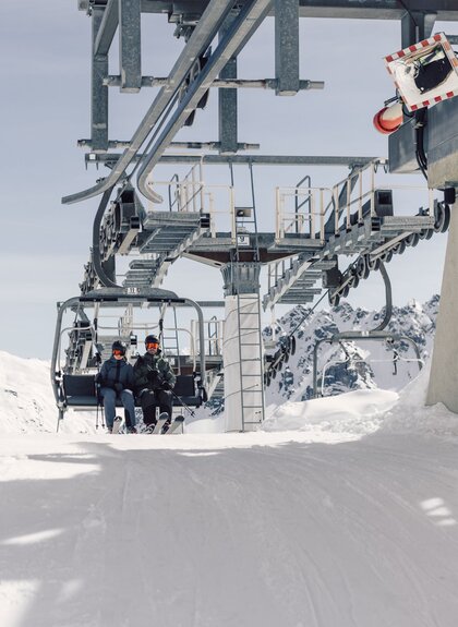Zwei Skifahrer erreichen mit dem Sessellift die Bergstation der Gargellner Bergbahnen. Ein Mitarbeiter überwacht den Ausstieg bei strahlendem Sonnenschein und verschneiter Bergkulisse. | © Gargellner Bergbahnen GmbH & Co KG, Daniel Zangerl