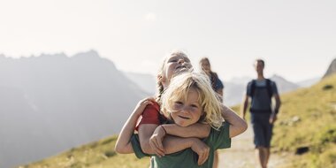 Zwei Kinder umarmen sich lachend auf einem sonnigen Wanderweg in Gargellen. Im Hintergrund folgen Erwachsene, während die umliegenden Berge in sanftem Licht erstrahlen. | © Gargellner Bergbahnen, Daniel Zangerl