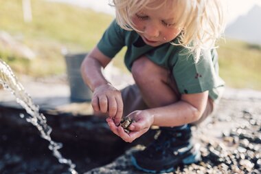 Ein kleiner blonger Junge hockt draußen an einer Wasserstelle und betrachtet konzentriert ein paar glänzende Goldsteine, die er in der Hand hält. Mit der einen Hand hält er die Fundstücke, mit der anderen untersucht er sie vorsichtig. Im Vordergrund fließt ein dünner Wasserstrahl aus einer Quelle. Der Junge trägt ein grünes T-Shirt, Shorts und robuste Wanderschuhe. Im Hintergrund ist ein Eimer zu sehen, und die Umgebung wirkt naturnah – mit Wiese, Felsen und Bergen im Gegenlicht. Die Szene strahlt Entdeckerfreude, Neugier und Naturverbundenheit aus. | © Gargellner Bergbahnen GmbH & Co KG, Daniel Zangerl