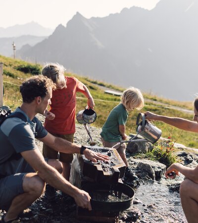 Eine Familie mit zwei Kindern wäscht gemeinsam Gold in den Bergen von Gargellen. Die Kinder gießen Wasser mit Kannen, während die Eltern beim Sieben helfen. Im Hintergrund sind grüne Wiesen und Berggipfel zu sehen. | © Gargellner Bergbahnen GmbH & Co KG, Daniel Zangerl