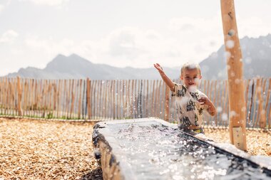 Junge spielt an einem Wasserspielelement auf dem Spielplatz am Schafberg, mit den Ritzensitzen und dem Schmalzberg im Hintergrund. | © Gargellner Bergbahnen GmbH & Co KG, Daniel Zangerl