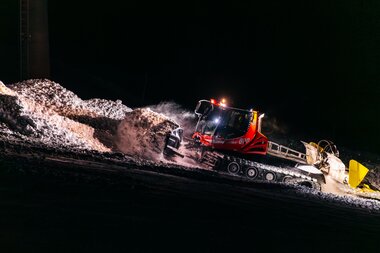 Eine rote Pistenraupe der Gargellner Bergbahnen schiebt in der Nacht frischen Schnee über die Piste. Im Schein der starken Scheinwerfer wirbelt der Schnee auf und glitzert im Licht – ein eindrucksvolles Bild der nächtlichen Präparationsarbeiten im Skigebiet Gargellen. | © Montafon Tourismus GmbH, Marie Schilcher