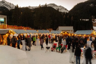 Belebter Wintermarkt in Gargellen mit beleuchteten Marktständen, Gästen im Schnee, Stehtischen und Liegestühlen vor verschneiten Bergen und dunklem Nadelwald in der Abenddämmerung. | © Montafon Tourismus, Marie Schilcher