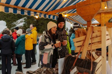 Zwei lächelnde Besucher mit Winterkleidung stehen an einem Marktstand mit handgefertigten Taschen und Waren; sie halten Becher in den Händen, im Hintergrund befinden sich weitere Marktbesucher unter blau-weiß gestreiften Marktdächern im Schnee. | © Montafon Tourismus, Marie Schilcher