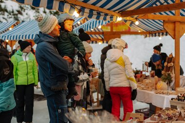Ein Erwachsener hält ein Kind auf dem Arm, während sie gemeinsam einen Marktstand mit handgefertigten Holzprodukten besuchen; rundherum stehen weitere Besucher in Winterkleidung unter blau-weiß gestreiften Marktdächern, warme Lichter beleuchten den verschneiten Wintermarkt. | © Montafon Tourismus, Marie Schilcher