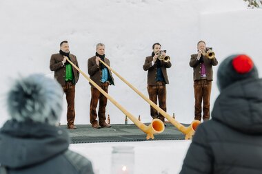 Vier Männer in traditioneller Tracht stehen auf einer kleinen Bühne im Schnee; zwei spielen Alphörner, zwei weitere Blasinstrumente, während Zuschauer im Vordergrund zuhören. | © Montafon Tourismus, Marie Schilcher