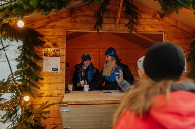Zwei Mitarbeiterinnen der Gargellner Bergbahnen stehen hinter einer hölzernen Ausschankhütte auf dem Weihnachtsmarkt in Gargellen und servieren Heißgetränke an Besucher. Die Hütte ist mit Tannenzweigen und Lichterketten dekoriert, im Vordergrund spricht eine Besucherin mit den Mitarbeiterinnen. | © Montafon Tourismus, Marie Schilcher