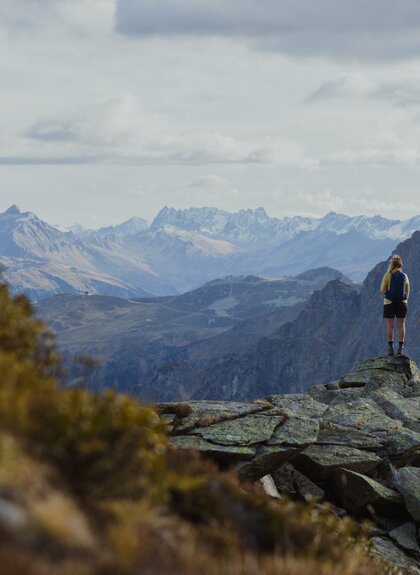 Zwei Wandernde stehen auf einem Felsen hoch über Gargellen und genießen den Blick auf die umliegenden Berge. Der Herbst taucht die Landschaft in sanfte Farben, während die schneebedeckten Gipfel in der Ferne leuchten – ein Moment der Ruhe und Weite in Gargellen. | © Gargellner Bergbahnen GmbH & Co KG, Alexander Furchs 