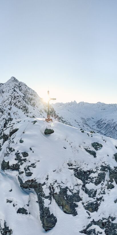 Verschneiter Berggipfel mit Gipfelkreuz im Sonnenlicht, umgeben von einer beeindruckenden winterlichen Berglandschaft im Montafon. | © Gargellner Bergbahnen GmbH & Co KG, Daniel Zangerl
