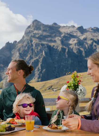 Eine Familie frühstückt auf einer Sonnenterrasse mit Blick auf die Berge von Gargellen. Eltern und Kinder sitzen an einem Holztisch, essen und lachen gemeinsam. Im Hintergrund ragt ein markanter Felsgipfel in den blauen Himmel, während andere Gäste auf der Terrasse sitzen. | © Gargellner Bergbahnen GmbH & Co KG, Alexander Fuchs