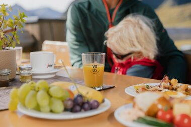 Ein Glas Orangensaft steht auf einem Holztisch neben Tellern mit frischem Obst, Brot und Frühstücksspeisen. Im Hintergrund sitzt ein Erwachsener mit einem Kind auf dem Schoß, im weichen Sonnenlicht einer Bergterrasse. | © Gargellner Bergbahnen GmbH & Co KG, Alexander Fuchs