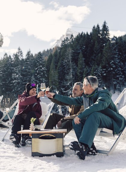 Drei Personen sitzen auf Liegestühlen im Schnee vor der Schirmbar in Gargellen und stoßen mit Getränken an; im Hintergrund sind verschneite Wälder, eine Gondelbahn und alpine Landschaft zu sehen. | © Gargellner Bergbahnen GmbH & Co KG, Daniel Zangerl