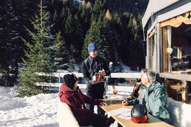 Eine Servicekraft serviert Getränke an vier sitzende Personen an einem Tisch im Schnee vor der Schirmbar in Gargellen; auf dem Tisch stehen Gläser, ein Skihelm liegt daneben, im Hintergrund sind verschneite Bäume und Skiausrüstung zu sehen. | © Gargellner Bergbahnen GmbH & Co KG, Daniel Zangerl