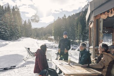 Ein Mitarbeiter serviert Getränke an einen Tisch mit vier Gästen im Schnee vor der Schirmbar in Gargellen; die Personen sitzen und stehen in Winterkleidung, im Hintergrund sind eine Gondelbahn, verschneite Bäume und Skipisten zu sehen. | © Gargellner Bergbahnen GmbH & Co KG, Daniel Zangerl