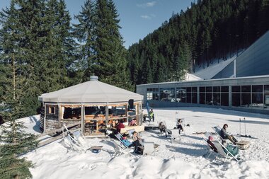 Gäste entspannen auf Liegestühlen im Schnee vor einem runden Bergrestaurant in Gargellen. Im Hintergrund sind verschneite Hänge, ein moderner Gebäudekomplex und dichter Nadelwald bei sonnigem Winterwetter zu sehen. | © Gargellner Bergbahnen GmbH & Co KG, Daniel Zangerl