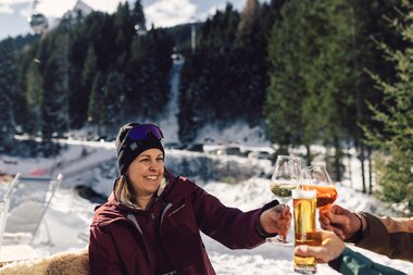Eine Frau in Winterbekleidung stößt im Schnee vor der Schirmbar in Gargellen mit zwei weiteren Personen an; sie hält ein Weinglas, daneben sind ein Bierglas und ein weiteres Getränk zu sehen, im Hintergrund verschneiter Wald und Skipiste. | © Gargellner Bergbahnen GmbH & Co KG, Daniel Zangerl