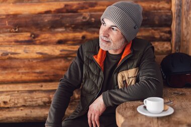 Ein Mann sitzt entspannt in der Schirmbar in Gargellen an einem Holztisch mit einer Tasse Kaffee und blickt nachdenklich zur Seite; im Hintergrund ist eine Holzverkleidung der Hütte zu sehen. | © Gargellner Bergbahnen GmbH & Co KG, Daniel Zangerl