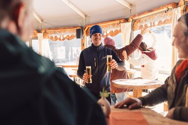 Mehrere Gäste stehen in einer gemütlichen Schirmbar in Gargellen. Eine Person hält zwei gefüllte Biergläser in den Händen, während andere im Hintergrund lachen, an Stehtischen stehen und miteinander sprechen. Warmes Licht und Holzelemente prägen die Atmosphäre. | © Gargellner Bergbahnen GmbH & Co KG, Daniel Zangerl