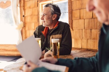 Ein Mann sitzt in einer gemütlichen Skihütte aus Holz an einem Tisch mit zwei Gläsern Bier und blickt entspannt aus dem Fenster; im Vordergrund halten weitere Gäste eine Speisekarte. | © Gargellner Bergbahnen GmbH & Co KG, Daniel Zangerl