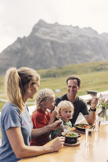 Eine Familie mit zwei Kindern sitzt an einem Holztisch auf einer Sonnenterrasse und genießt ihr Essen mit Blick auf die Berge von Gargellen. Im Hintergrund ragt ein markanter Felsen auf, während alle lächeln und die Mahlzeit gemeinsam genießen. Auf dem Tisch stehen Schüsseln mit bunten Gerichten und Gläser mit Wasser. | © Gargellner Bergbahnen GmbH & Co KG, Daniel Zangerl