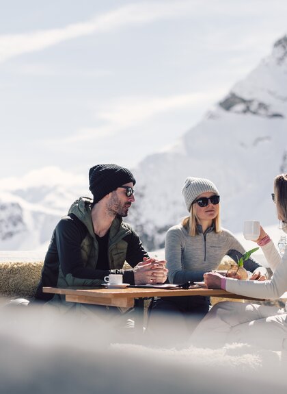 Drei Personen sitzen auf einer mit Fellen belegten Bank an einem Holztisch auf einer Sonnenterrasse in den Bergen. Sie tragen Winterkleidung mit Mützen und Sonnenbrillen und genießen Getränke in der Sonne. Im Hintergrund sind schneebedeckte Berge und ein blauer Himmel zu sehen. | © Gargellner Bergbahnen GmbH & Co KG, Daniel Zangerl