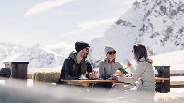 Drei Personen sitzen auf einer mit Fellen belegten Bank an einem Holztisch auf einer Sonnenterrasse in den Bergen. Sie tragen Winterkleidung mit Mützen und Sonnenbrillen und genießen Getränke in der Sonne. Im Hintergrund sind schneebedeckte Berge und ein blauer Himmel zu sehen. | © Gargellner Bergbahnen GmbH & Co KG, Daniel Zangerl