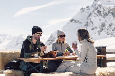 Drei Personen sitzen auf einer mit Fellen belegten Bank an einem Holztisch auf einer Sonnenterrasse in den Bergen. Sie tragen Winterkleidung mit Mützen und Sonnenbrillen und genießen Heißgetränke in der Sonne. Hinter ihnen sind schneebedeckte Gipfel und blauer Himmel zu sehen. | © Gargellner Bergbahnen GmbH & Co KG, Daniel Zangerl