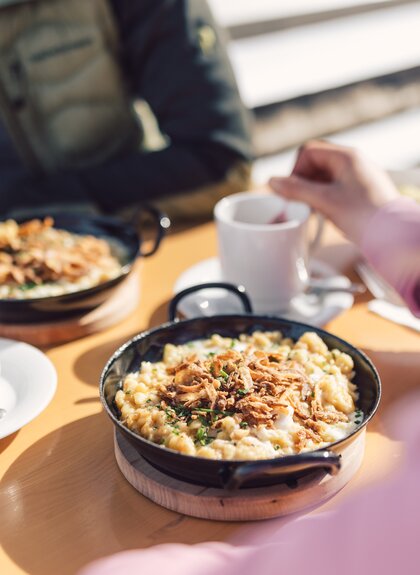 Zwei gusseiserne Pfannen mit frisch servierten Käsespätzle und Röstzwiebeln stehen auf einem Holztisch in der Sonne. Daneben halten zwei Personen weiße Tassen mit warmem Getränk. Die Szene spielt im Freien, das Licht ist warm und freundlich. | © Gargellner Bergbahnen GmbH & Co KG, Daniel Zangerl