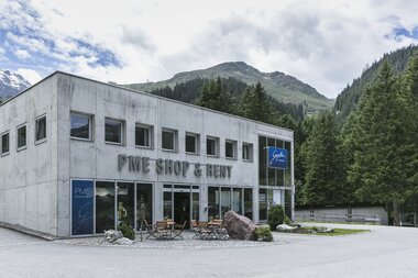 Das Gebäude des PME Shop & Rent in Gargellen steht vor einer Kulisse aus grünen Bergen und dichten Wäldern. Das zweistöckige Haus aus grauem Beton hat große Glasfronten im Erdgeschoss mit der Aufschrift „PME Shop & Rent“ sowie dem blauen Schild „Gargellen Montafon“. Vor dem Eingang stehen Tische und Stühle auf einer kleinen Terrasse. | © Gargellner Bergbahnen GmbH & Co KG, Christoph Schöch
