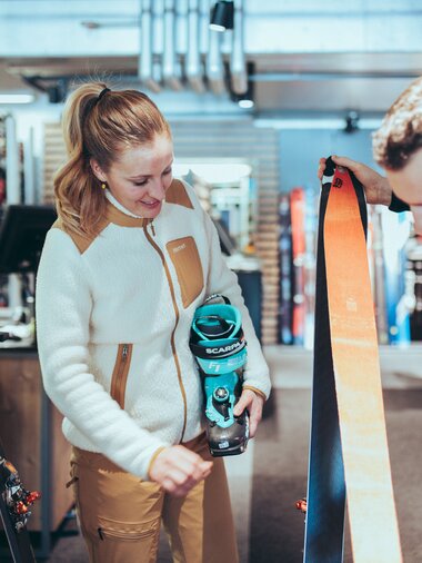 In einem Skiverleih berät ein Mitarbeiter eine Frau zur passenden Tourenausrüstung. Die Frau hält einen türkisfarbenen Skitourenschuh der Marke Scarpa, während der Mitarbeiter ein Paar Ski mit orangefarbenen Fellen zeigt. Im Hintergrund sind weitere Ski und Ausrüstungsgegenstände zu sehen. | © Gargellner Bergbahnen GmbH & Co KG, Christoph Schöch