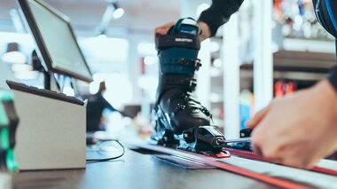 Eine Person stellt mit einem Schraubenzieher die Bindung eines Skis ein, in dem ein schwarzer Skischuh der Marke Salomon eingespannt ist. Der Ski liegt auf einer Werkbank in einem modernen Skiverleih. Im Hintergrund sind Arbeitsgeräte und Regale leicht unscharf zu erkennen. | © Gargellner Bergbahnen GmbH & Co KG, Christoph Schöch
