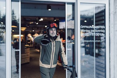 Ein Wintersportgast mit Helm und Skibrille tritt durch den Eingang eines Sportshops und hält Skier in der Hand; hinter ihm ist der Innenbereich des Geschäfts sichtbar, an der Glastür stehen die Öffnungszeiten. | © Gargellner Bergbahnen GmbH & Co KG, Daniel Zangerl