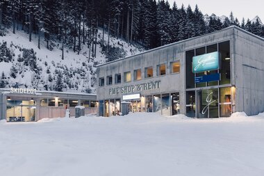 Außenansicht des Gebäudes „PME Shop & Rent“ mit angeschlossenem Skidepot im Skigebiet Gargellen; vor dem modernen Betonbau liegt Schnee, im Hintergrund ein bewaldeter, verschneiter Hang. | © Gargellner Bergbahnen GmbH & Co KG, Daniel Zangerl