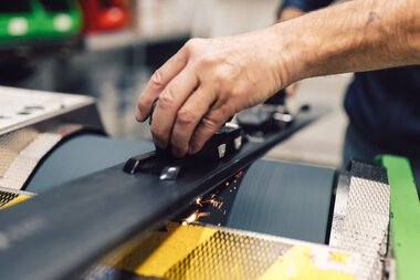 Nahaufnahme einer Hand, die einen Ski während des Kantenschleifens im Skiservice des PME Shop & Rent führt; der Ski liegt auf der Schleiffläche, an der Metallkante entstehen Funken. | © Gargellner Bergbahnen GmbH & Co KG, Daniel Zangerl