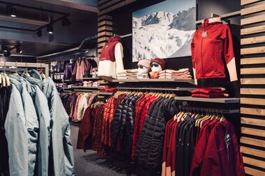 Übersicht über den Innenbereich des PME Shop & Rent mit aufgehängter Wintersportbekleidung in verschiedenen Farben; Jacken, Pullover und Accessoires sind geordnet an Kleiderstangen und Regalen präsentiert. | © Gargellner Bergbahnen GmbH & Co KG, Daniel Zangerl