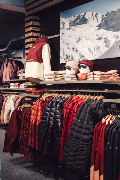 Übersicht über den Innenbereich des PME Shop & Rent mit aufgehängter Wintersportbekleidung in verschiedenen Farben; Jacken, Pullover und Accessoires sind geordnet an Kleiderstangen und Regalen präsentiert. | © Gargellner Bergbahnen GmbH & Co KG, Daniel Zangerl