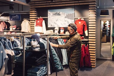 Ein Kunde steht im Innenbereich des PME Shop & Rent und betrachtet Winterjacken an einem Kleiderständer; rundherum sind weitere Wintersportbekleidung, Accessoires und Präsentationsflächen im Shop zu sehen. | © Gargellner Bergbahnen GmbH & Co KG, Daniel Zangerl