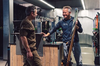 Ein Mitarbeiter des PME Shop & Rent steht im Verleihbereich neben einem Kunden und hält ein Paar Skier in der Hand; beide Personen führen ein Beratungsgespräch, im Hintergrund sind aufgestellte Skier zu sehen. | © Gargellner Bergbahnen GmbH & Co KG, Daniel Zangerl