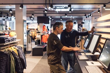 Zwei Personen stehen im Innenbereich des PME Shop & Rent an einer Service-Theke und bedienen Bildschirme; ein Mitarbeiter unterstützt einen Kunden bei der Datenerfassung, im Hintergrund sind Verkaufsflächen mit Wintersportbekleidung zu sehen. | © Gargellner Bergbahnen GmbH & Co KG, Daniel Zangerl