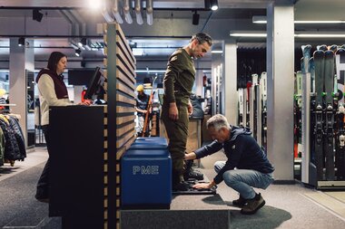 Ein Mitarbeiter passt einem Kunden im Innenbereich des PME Shop & Rent die Skischuhe an, während der Kunde auf einer kleinen Plattform steht; im Hintergrund sind der Kassenbereich, weitere Kunden und aufgestellte Skier zu sehen. | © Gargellner Bergbahnen GmbH & Co KG, Daniel Zangerl
